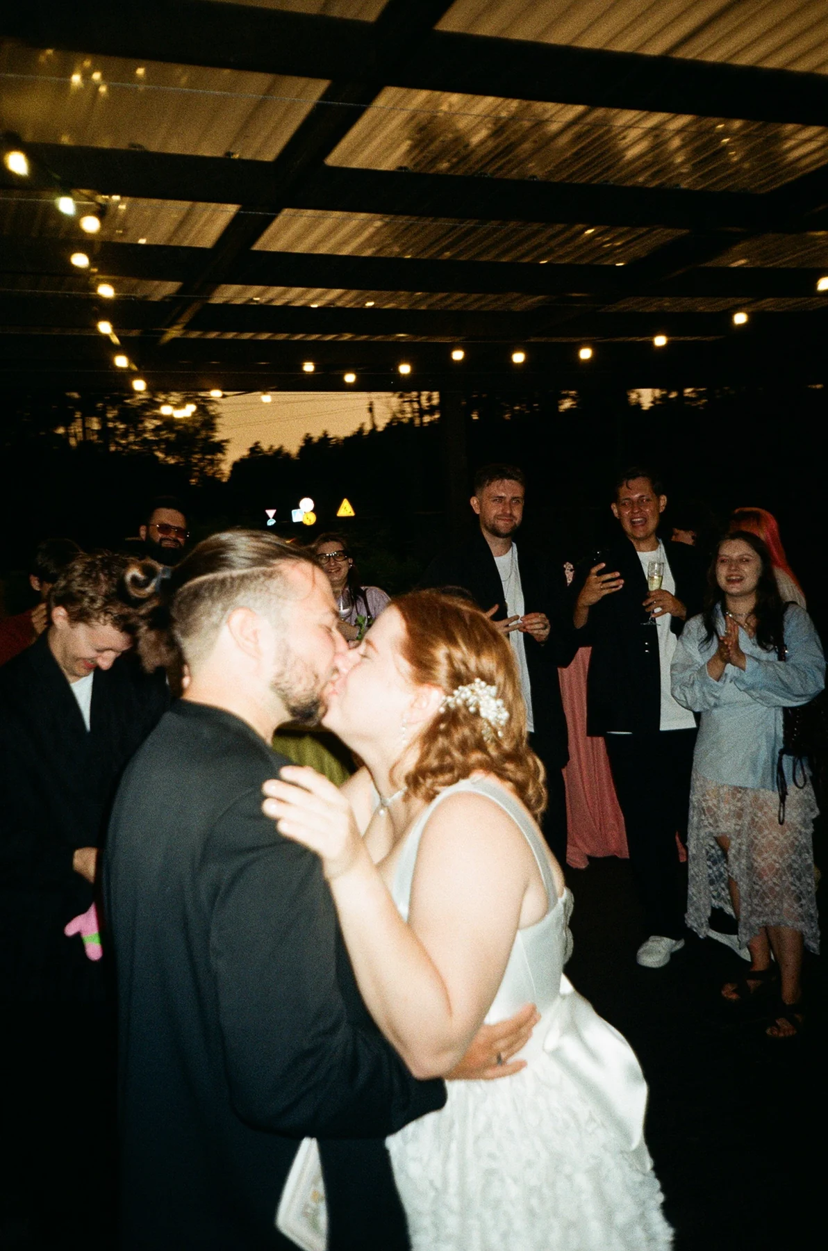 Newlyweds kissing among guests at a dark reception in a direct-flash film photograph